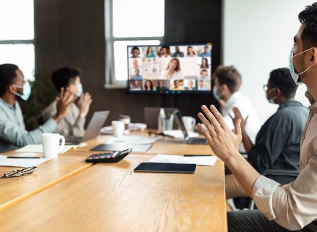 Diverse businesspeople making online video call to employees Videoconferencing Concept. Diverse group of workers in face masks making video call with colleagues, sitting at table in boardroom, waving at screen at office, talking on web with remote employees